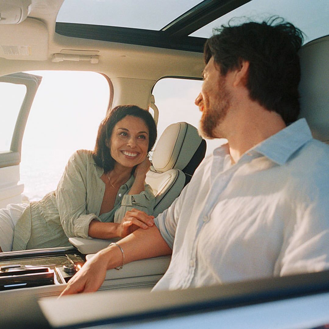 A smiling couple inside a car with a large sunroof and the door open, possibly at the beach, wearing casual linen clothes during daytime.