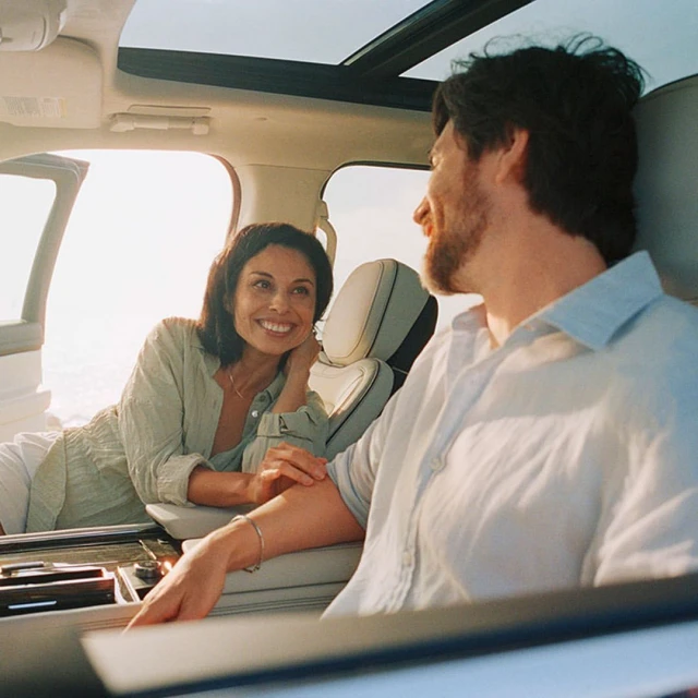A smiling couple inside a car with a large sunroof and the door open, possibly at the beach, wearing casual linen clothes during daytime.