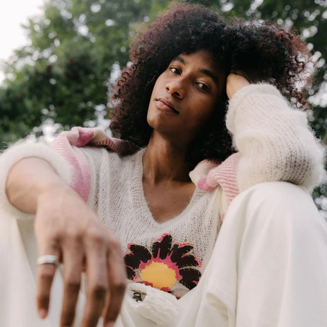 A young adult with curly hair wears a white sweater with a flower design and white pants, posing outdoors against a blurred green background.