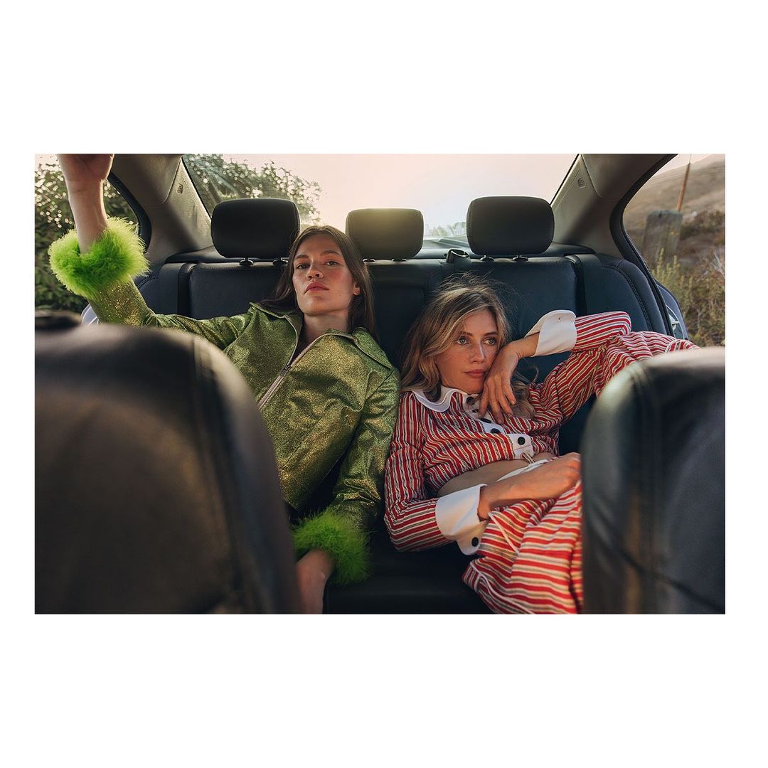 Two women in a car, one in a green jacket with fur trim and the other in red and white striped pajamas, are relaxing in the backseat.
