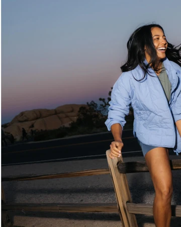 Smiling woman in blue quilted jacket leaning against a wooden fence at dusk with desert landscape behind