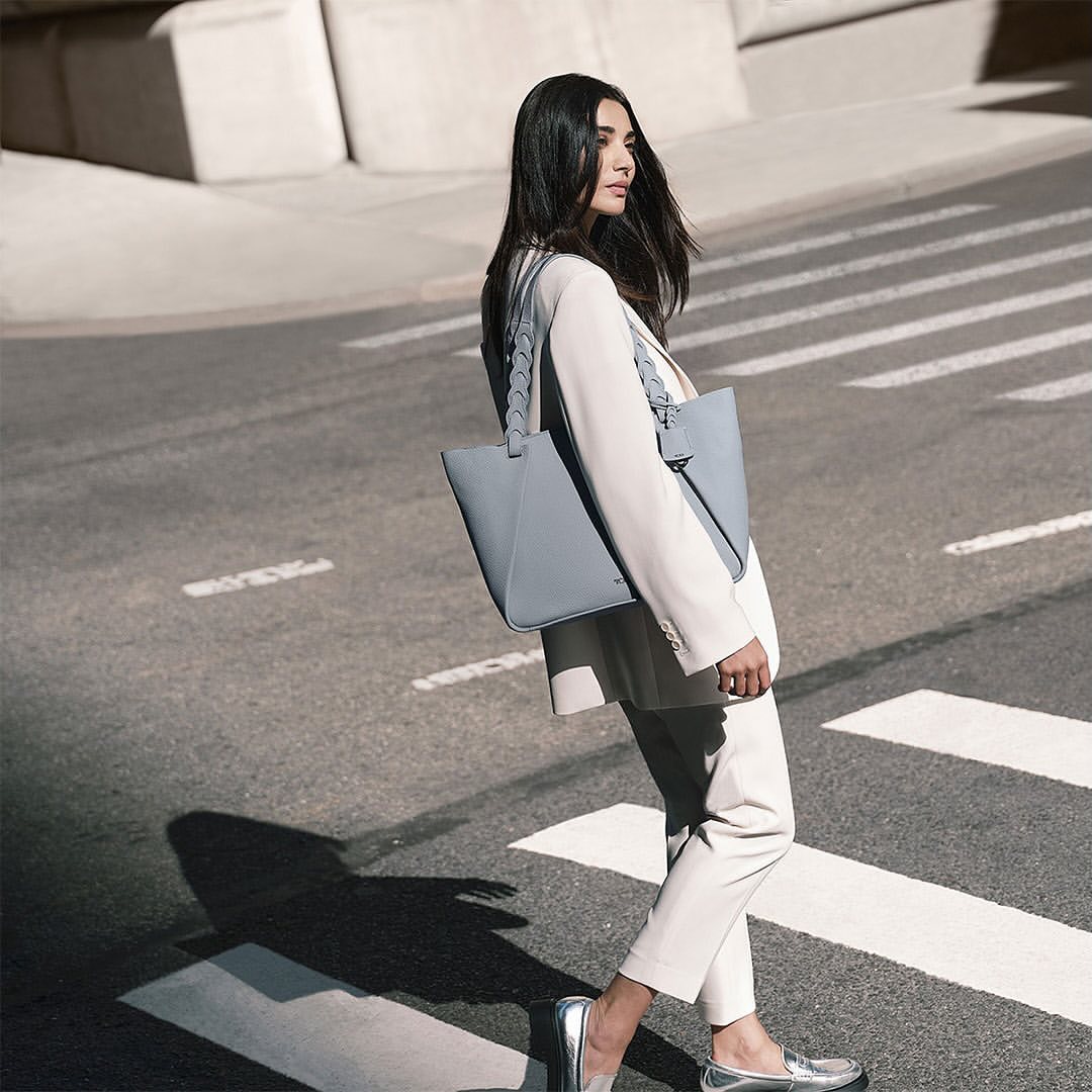 Woman in a white suit with a grey tote bag crossing a city street on a sunny day, modeling a fashion style.