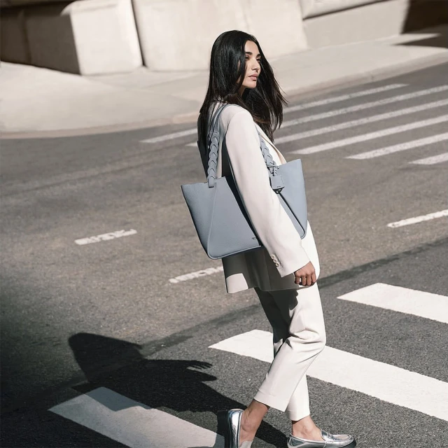 Woman in a white suit with a grey tote bag crossing a city street on a sunny day, modeling a fashion style.