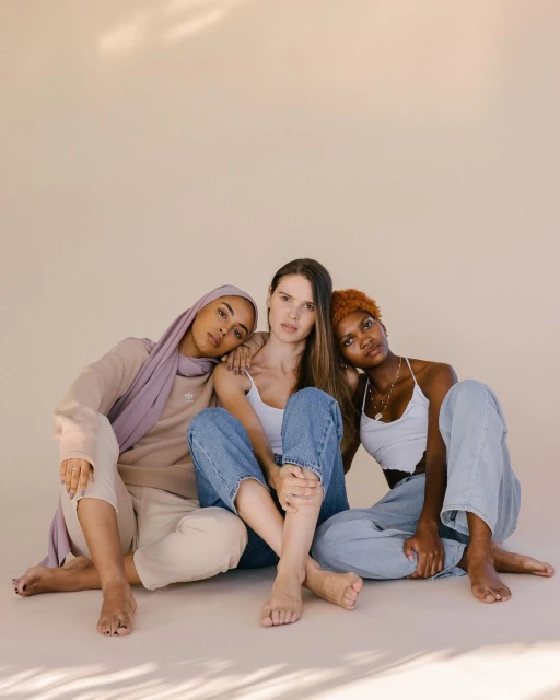Three women of diverse ethnicities sit together, wearing casual tops and jeans, creating a sense of unity in a photo studio setting.