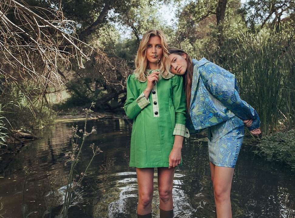 Two young adult women pose in retro-style dresses and boots, one in green holding a flower, in a nature scene with water.
