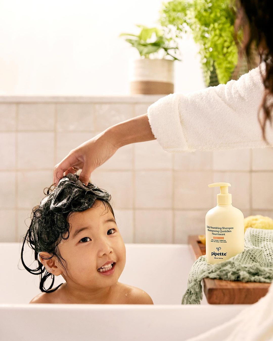 Young child with soapy hair is getting a shampoo in a white bathtub. A bottle of baby shampoo sits on a wooden tray.