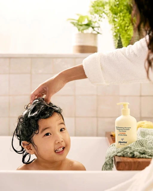 Young child with soapy hair is getting a shampoo in a white bathtub. A bottle of baby shampoo sits on a wooden tray.