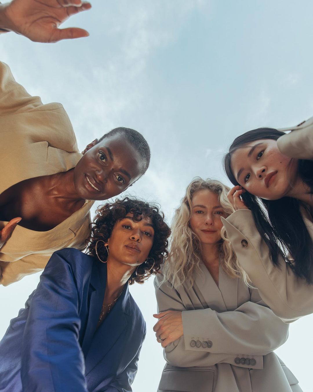 Four diverse women in blazers pose in a circle, shot from a low angle against a cloudy sky. Fashion-forward, stylish, and confident.