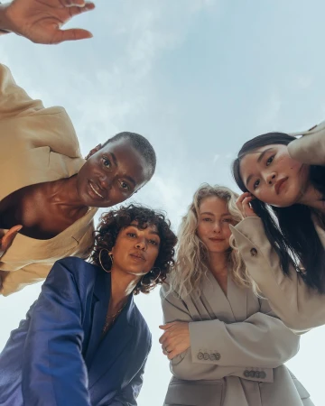 Four diverse women in blazers pose in a circle, shot from a low angle against a cloudy sky. Fashion-forward, stylish, and confident.