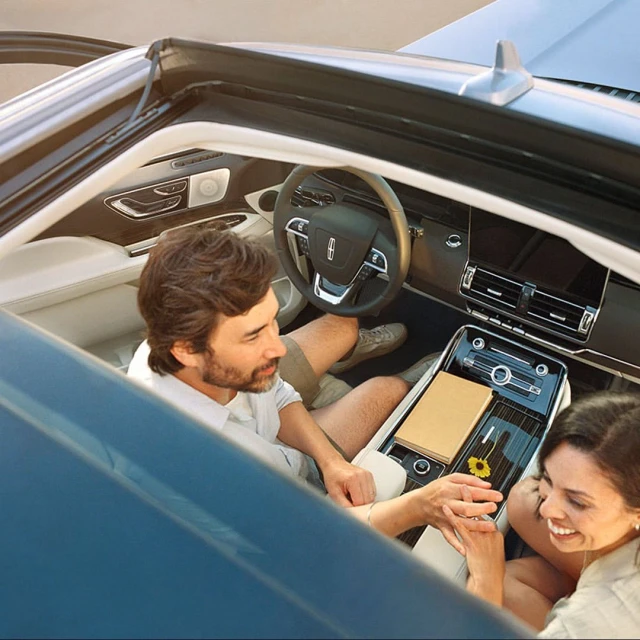 A couple sits inside a convertible Lincoln car with a tan notebook and flower on the console, capturing a moment of automotive elegance.
