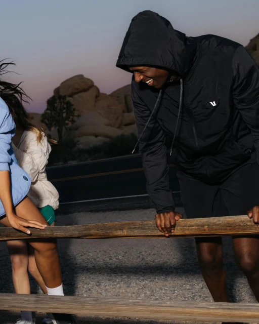 Two women and a man in workout gear on a dirt trail with scrub and rocks in the background, laughing while leaning on a wood railing.