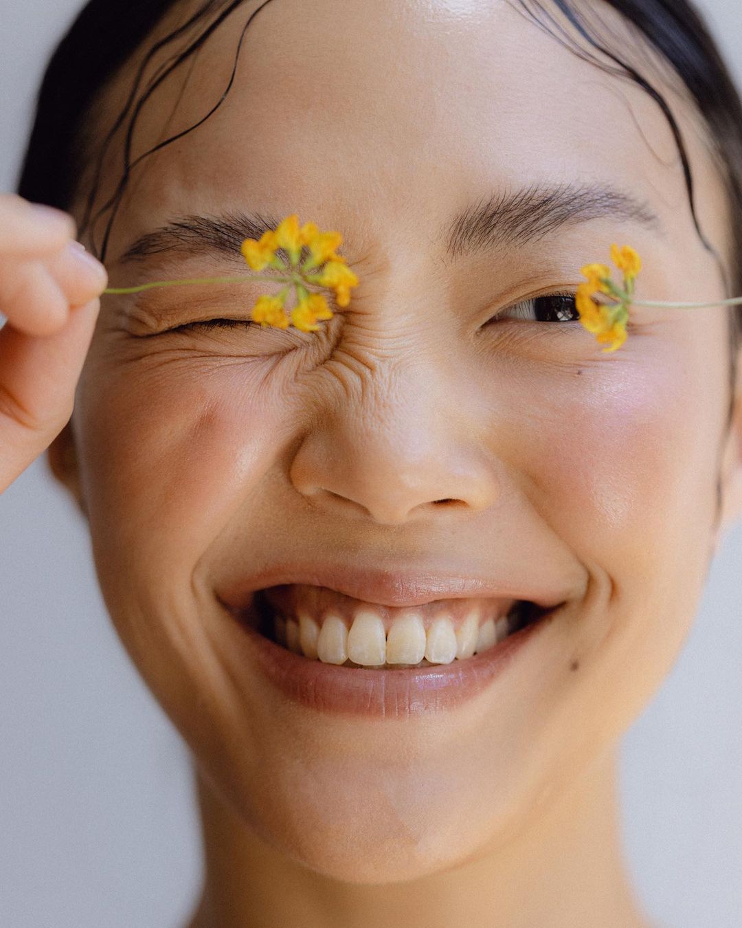 Portrait of smiling Asian woman with yellow flowers covering her eyes. Wet hair and glowing skin against a simple white background.