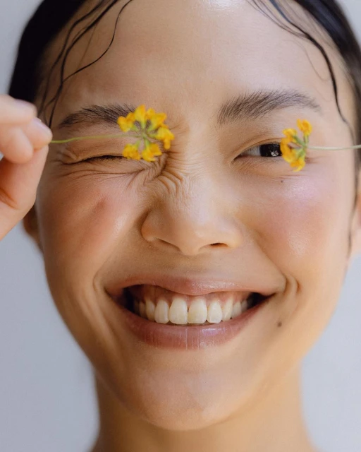 Portrait of smiling Asian woman with yellow flowers covering her eyes. Wet hair and glowing skin against a simple white background.