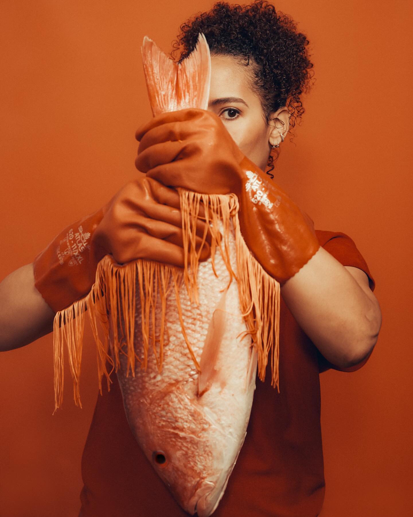 A woman wearing orange gloves holds a large fish against an orange background, partially obscuring her face in a studio shot.