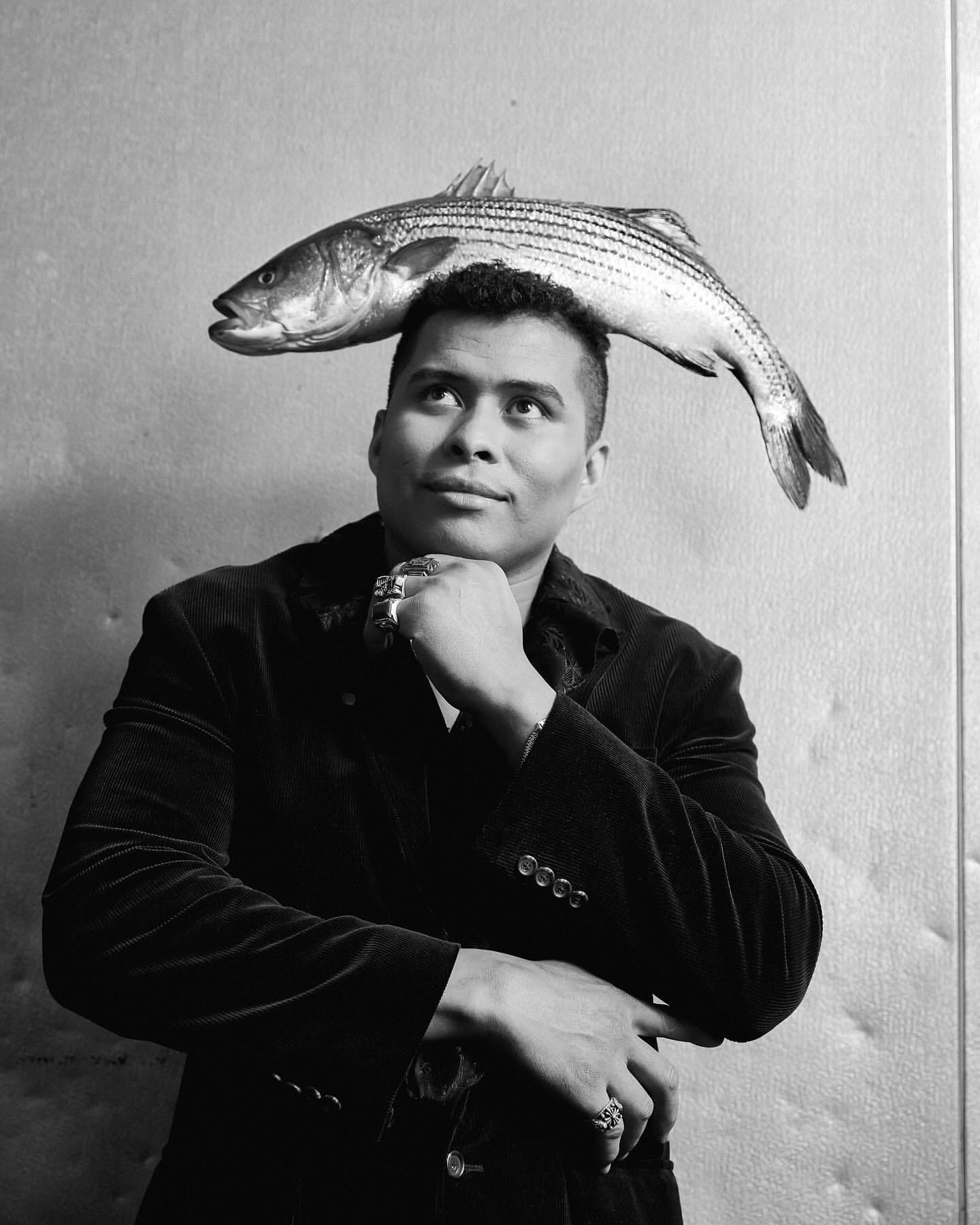 Portrait of an adult man wearing a jacket and rings, with a fresh fish balanced on top of his head in a black and white studio shot.