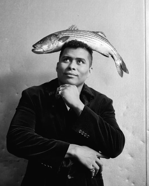 Portrait of an adult man wearing a jacket and rings, with a fresh fish balanced on top of his head in a black and white studio shot.