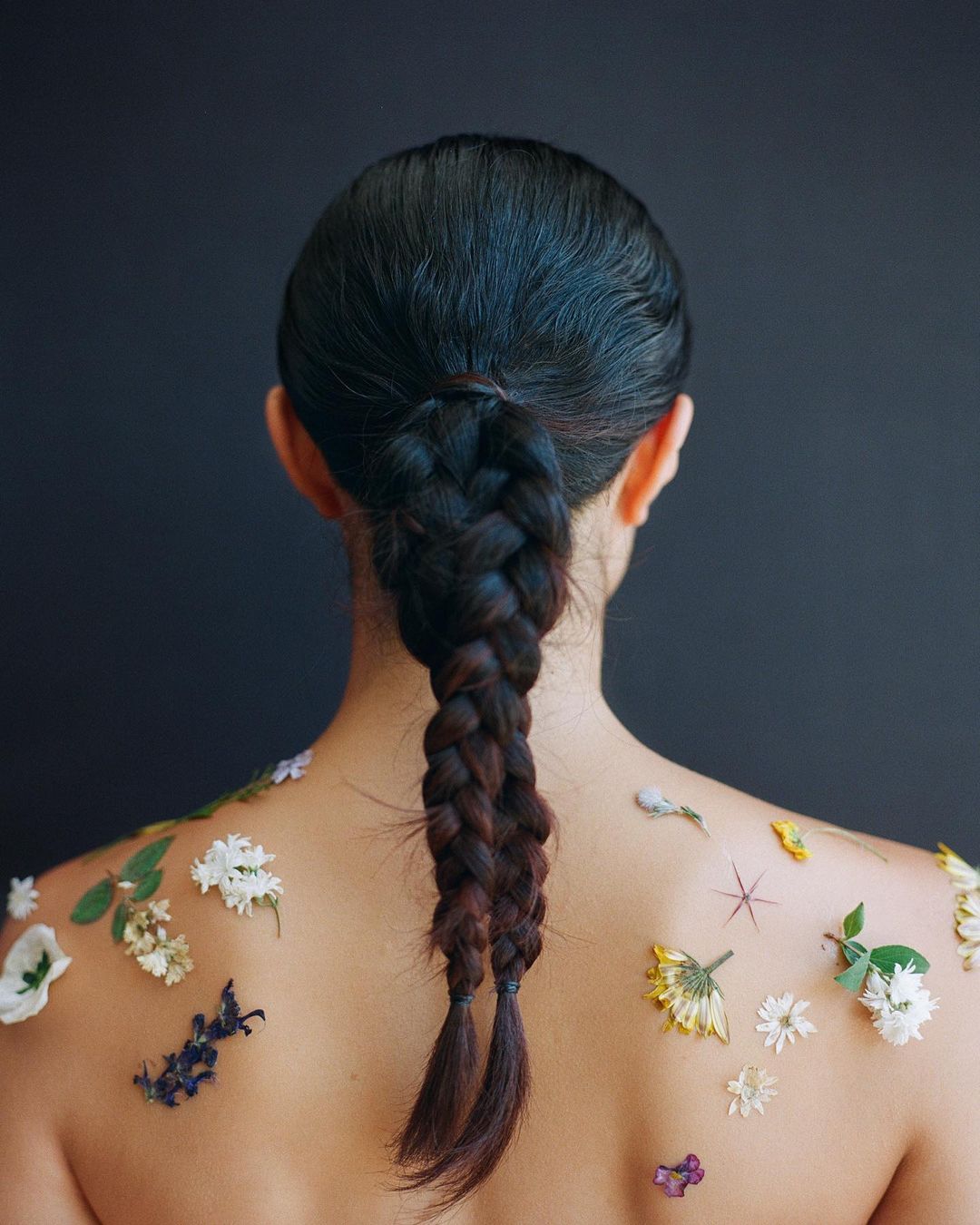 Back view of a person with dark hair in a braid, decorated with small white, yellow, and purple flowers on their shoulders and back.
