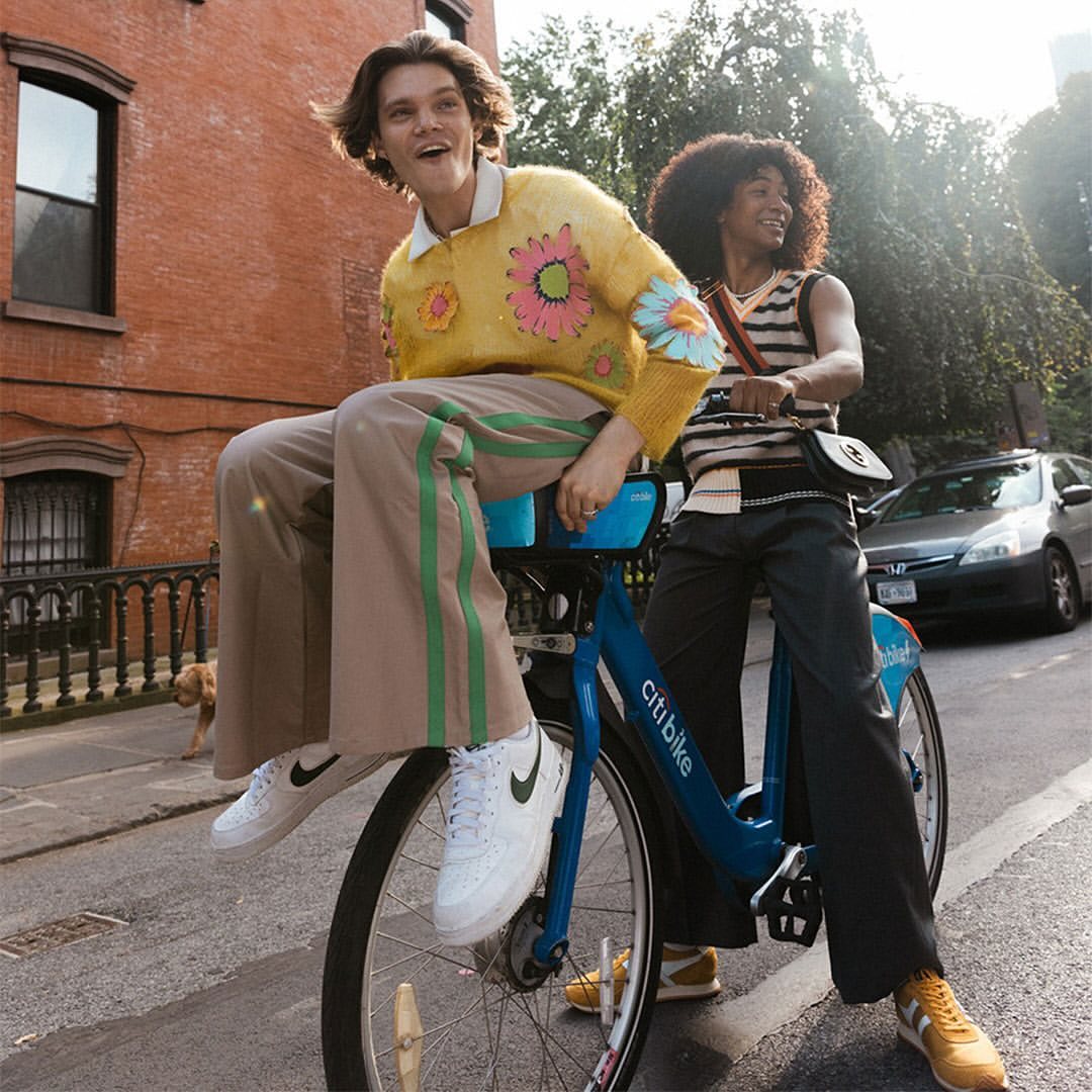 Two people ride a blue Citi Bike in a city street with a red brick building, car, and dog in the background on a sunny day.