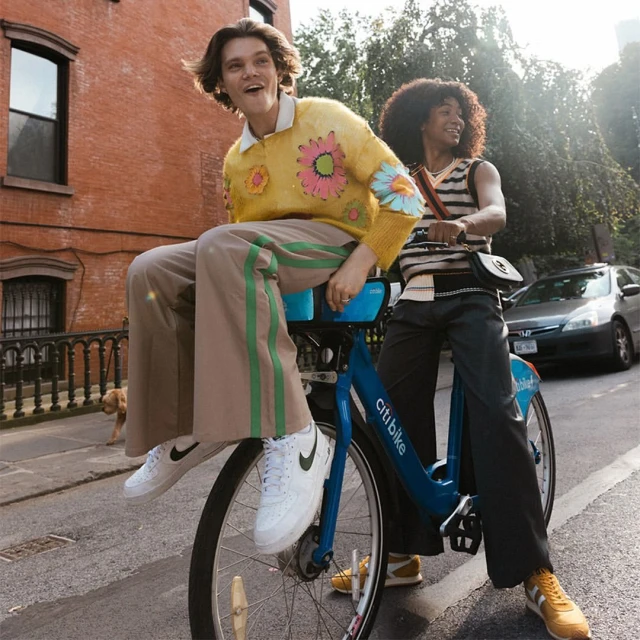Two people ride a blue Citi Bike in a city street with a red brick building, car, and dog in the background on a sunny day.