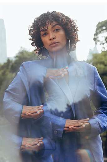 Portrait of an adult woman with curly hair, wearing a blue blazer, set in a park with trees and a tall building in the background.