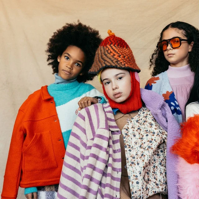Three young children pose indoors against a neutral backdrop wearing colorful sweaters, hats, and jackets in a fashion-forward style.