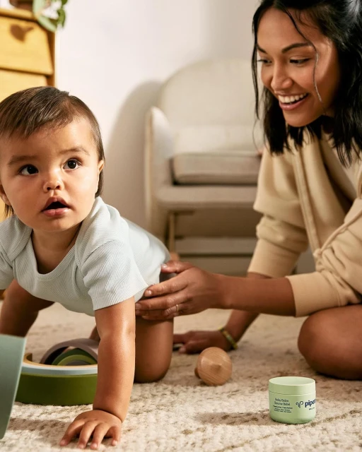 Happy baby crawling on the floor with their mother beside them. Cream jar of baby skincare product is visible. 