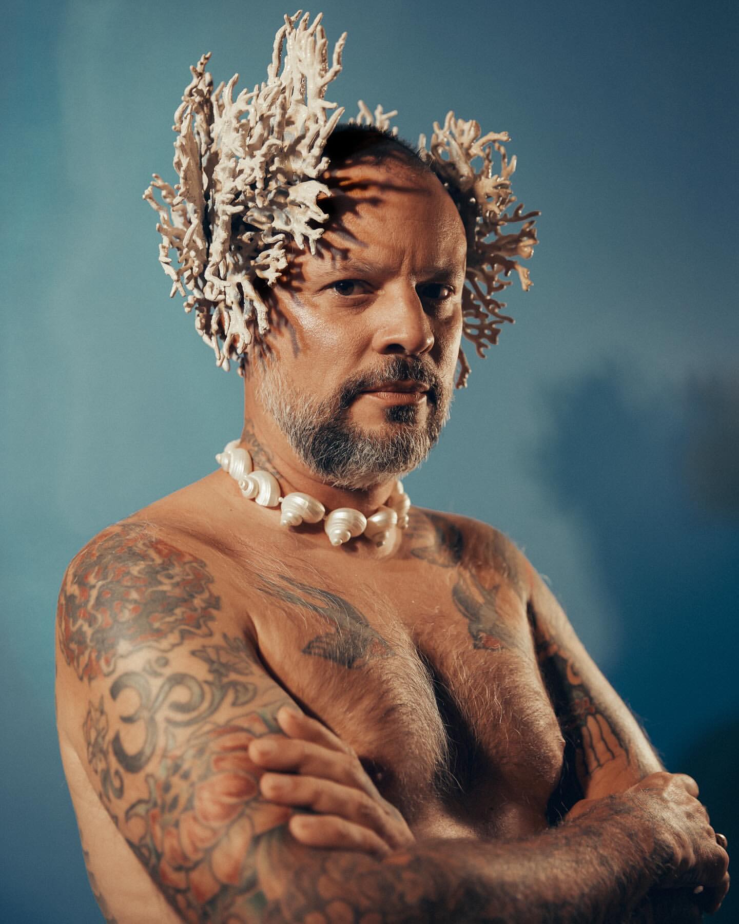 Portrait of a shirtless man with tattoos, a beard, a coral headpiece, and a shell necklace, set against a blue background with soft lighting.