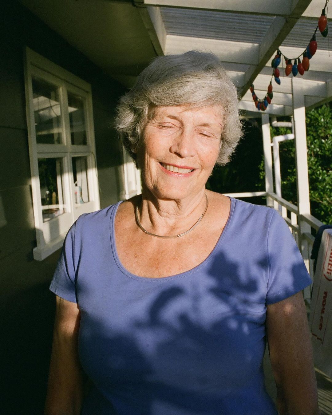 An elderly woman with short grey hair smiles with her eyes closed, wearing a blue t-shirt, outside on a sunny day with decorative lights.