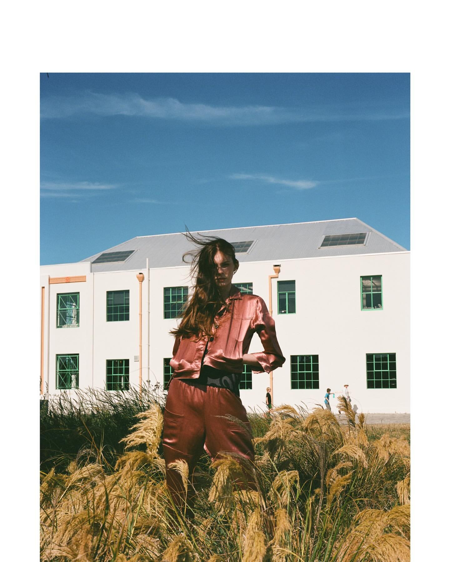 A woman in a satin outfit stands in a grassy field in front of a building. The shot is taken in daylight under a blue sky.