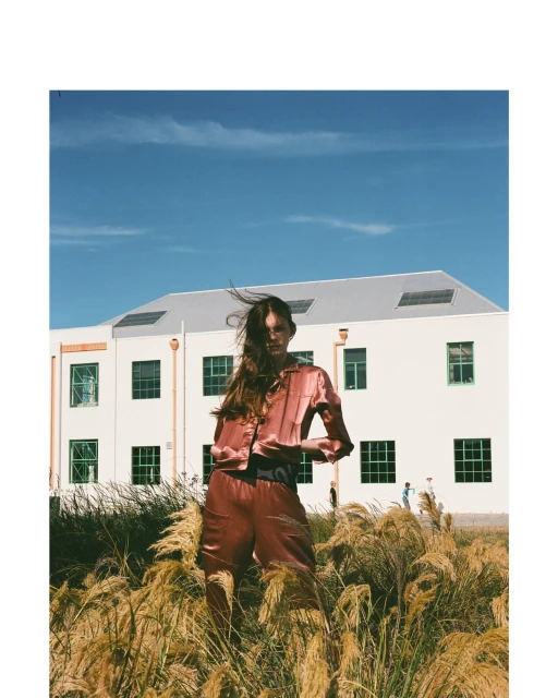 A woman in a satin outfit stands in a grassy field in front of a building. The shot is taken in daylight under a blue sky.