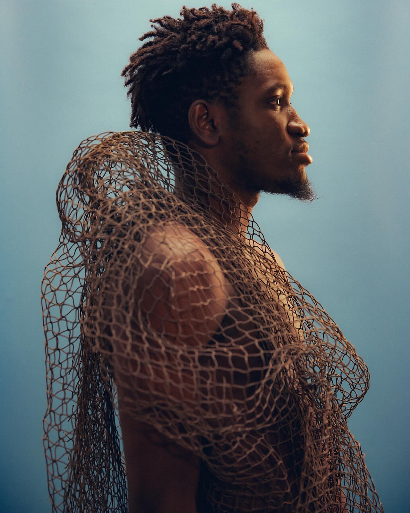 Profile portrait of an adult male model with dreadlocks, posing with a fishing net draped around his shoulders against a gradient blue background.