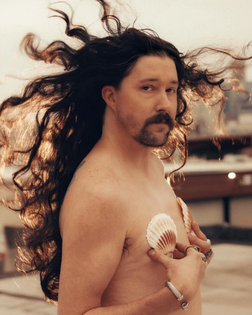 Man with long hair in artistic portrait. Upper body with multiple shell decorations. Outdoor scene. Rings and bracelet accessories.