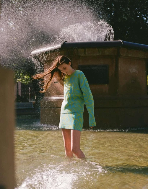 A young woman stands in a fountain wearing a green and blue patterned dress, with water splashing around her in a park setting.
