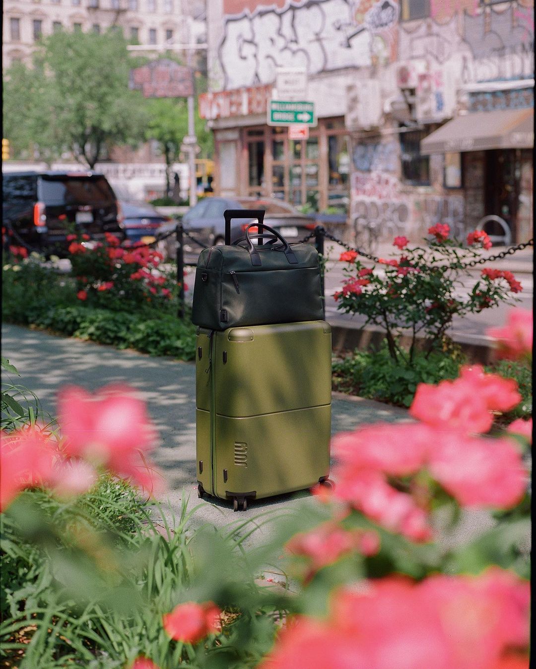 Olive green suitcase with a black duffel bag stacked on top, displayed outdoors with pink flowers and a building in the background.