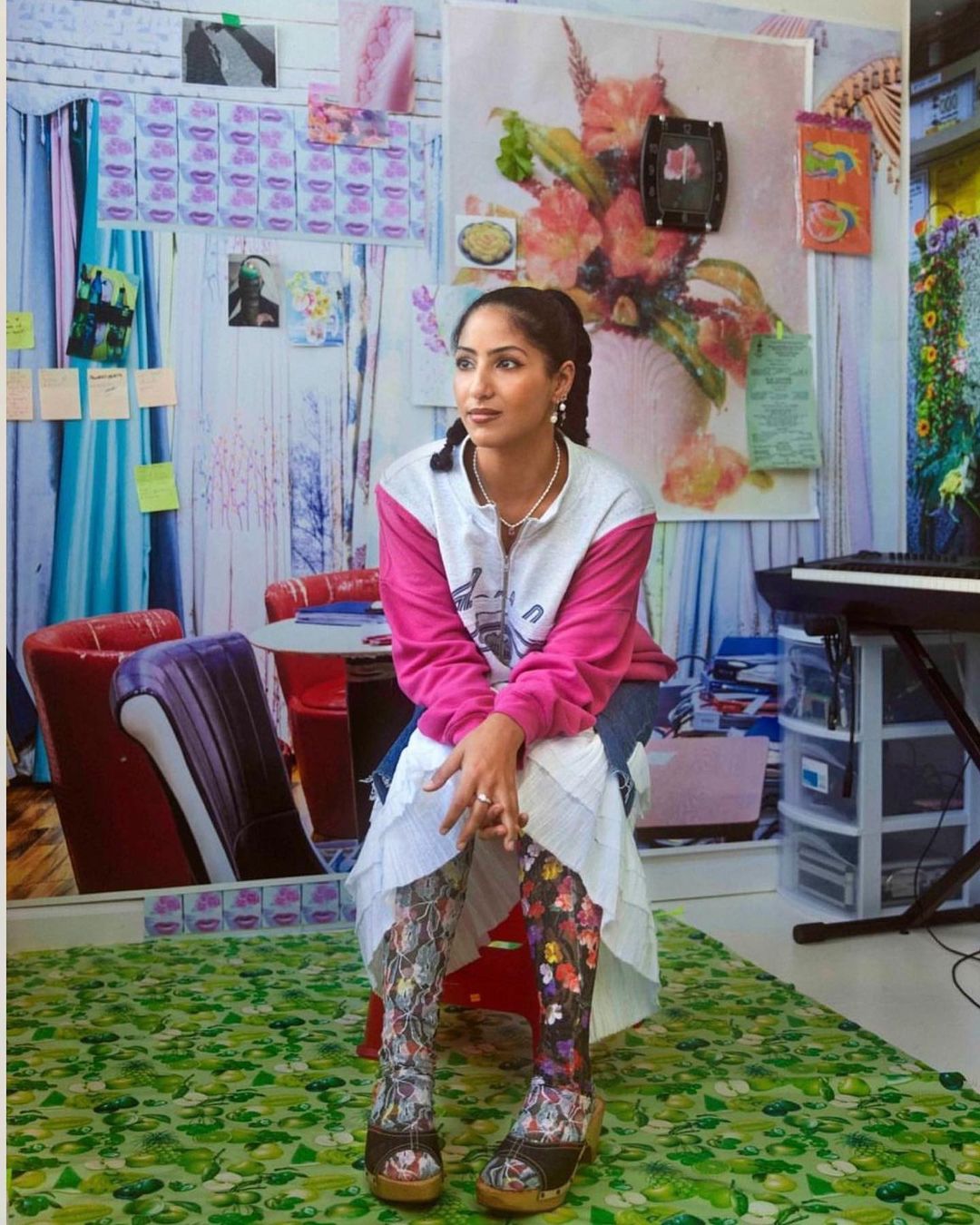 Woman in floral leggings sits in a room with wallpaper covered in art, images, and photos, plus a piano and colorful decor.