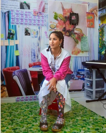 Woman in floral leggings sits in a room with wallpaper covered in art, images, and photos, plus a piano and colorful decor.