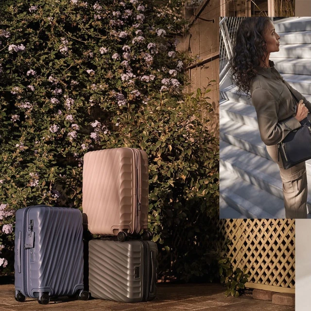 Three carry-on suitcases in blue, beige, and green sit in front of a flowering vine and a woman walks up stairs in the background.