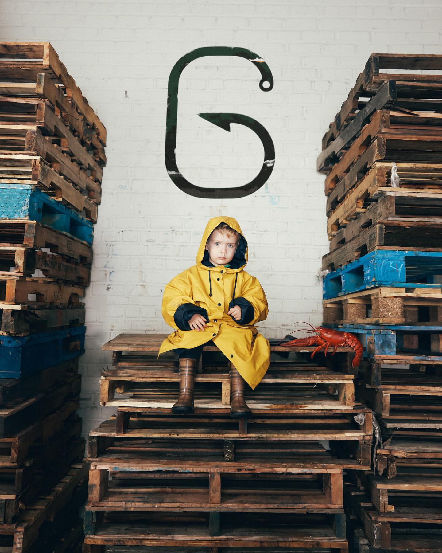 Young child in yellow raincoat sits on wooden pallets between stacks, with lobster and hook graphic on white brick wall background.
