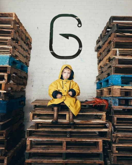 Young child in yellow raincoat sits on wooden pallets between stacks, with lobster and hook graphic on white brick wall background.