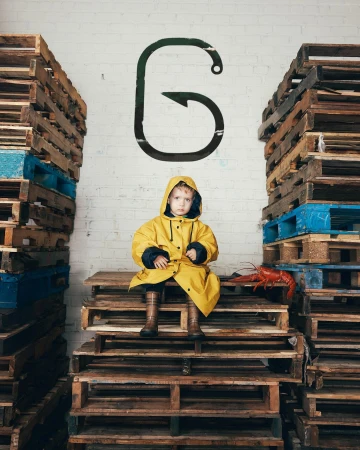 Young child in yellow raincoat sits on wooden pallets between stacks, with lobster and hook graphic on white brick wall background.