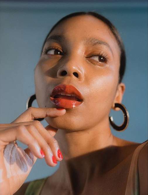 Close-up portrait of a Black woman with red lipstick and hoop earrings, looking away against a teal background with soft, diffused lighting.