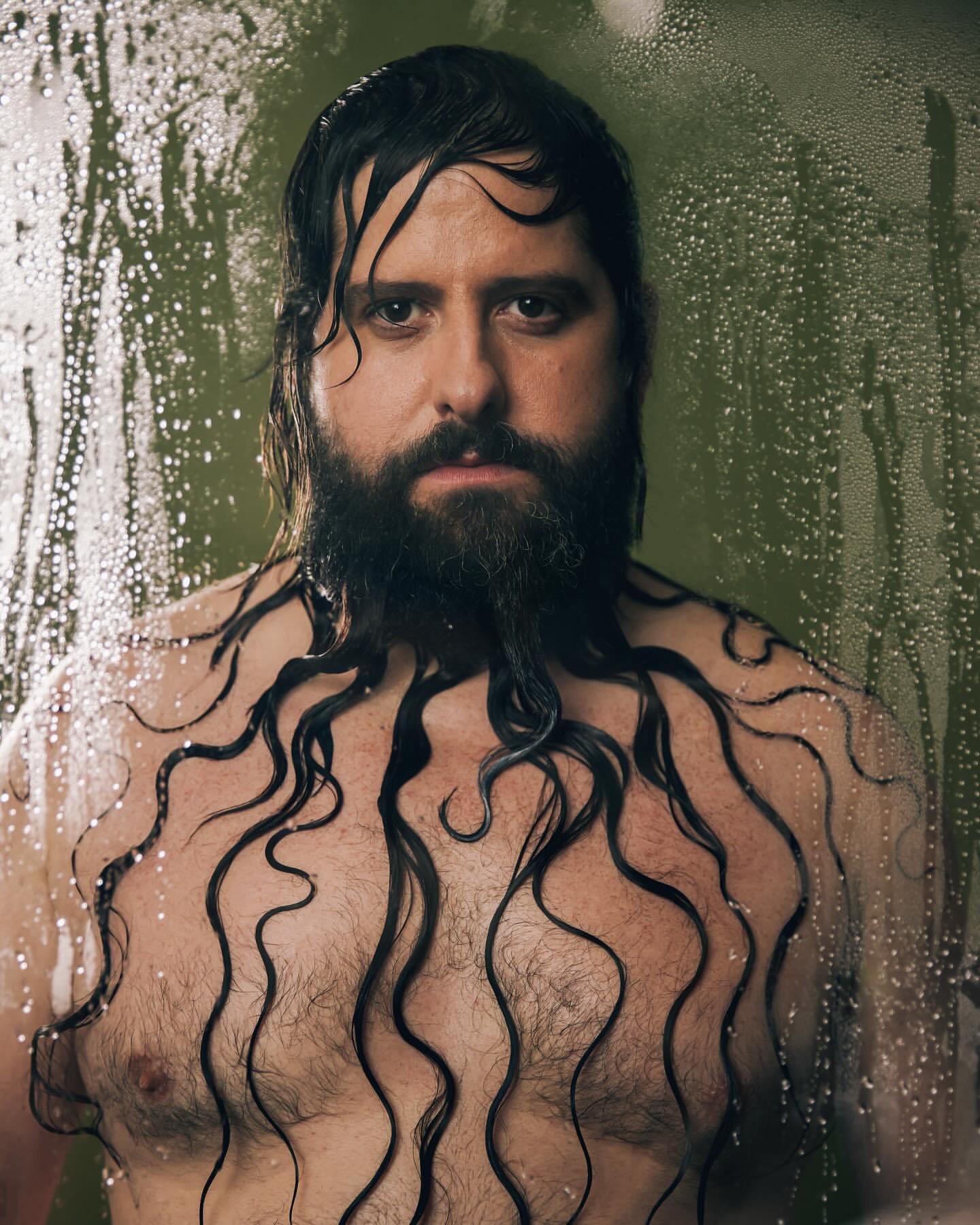Man with a beard stands in a shower with water droplets visible on the glass, the water streaming from his hair onto his chest.