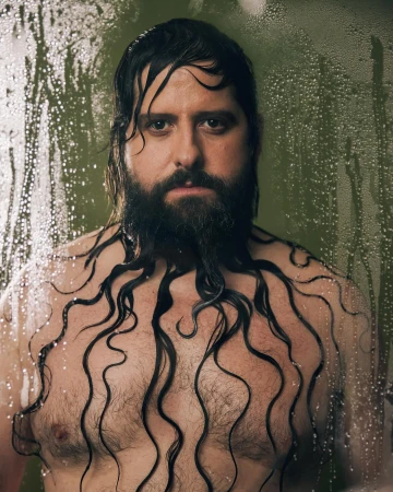 Man with a beard stands in a shower with water droplets visible on the glass, the water streaming from his hair onto his chest.