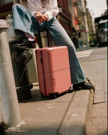 Person in wide-leg blue jeans and white shirt holding a pink suitcase on a city street curb in New York.