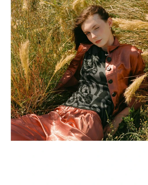 Woman in a brown jacket and pants lying in a field of tall grass, looking at the camera with a soft, natural light and editorial style.