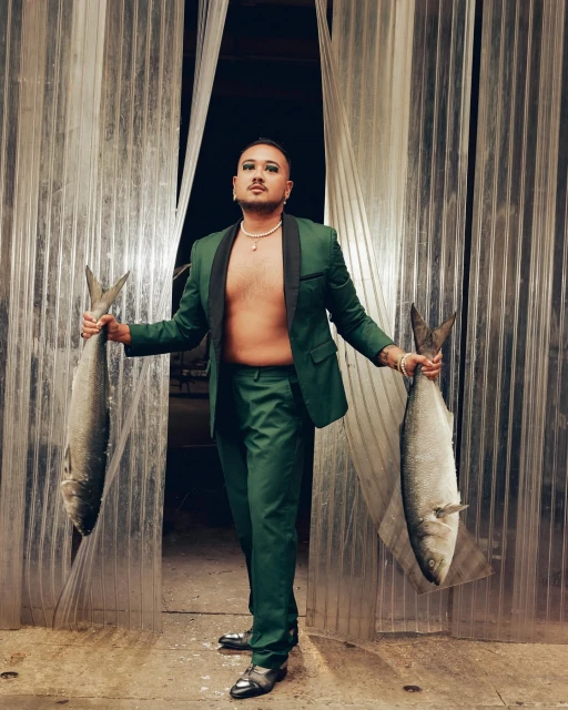 A person in a green suit stands in front of a translucent curtain holding large fish in each hand, creating a surreal fashion portrait.