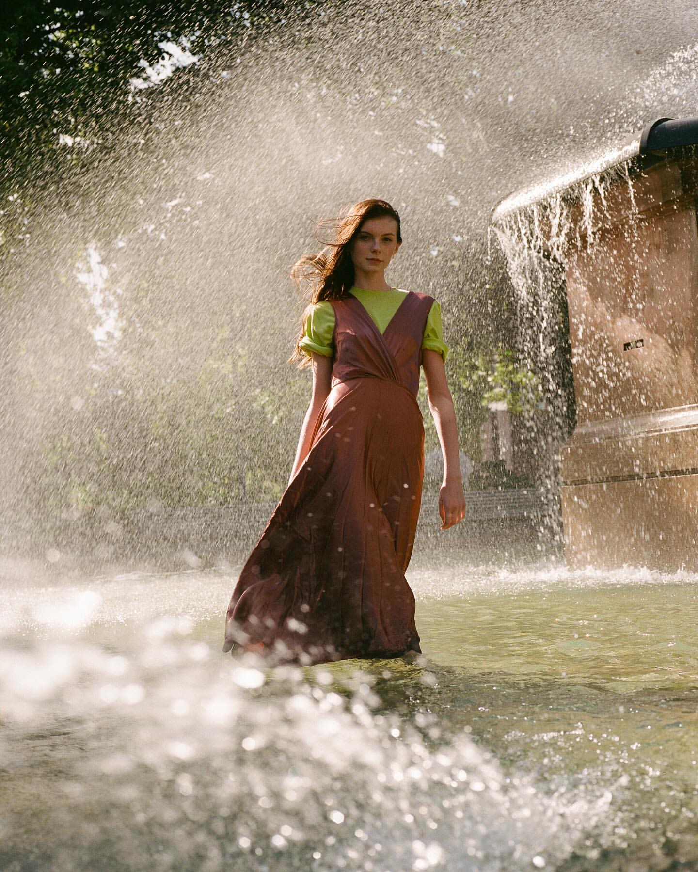 A woman stands in a fountain wearing a long brown dress with a green shirt, with water splashing around her on a sunny day.