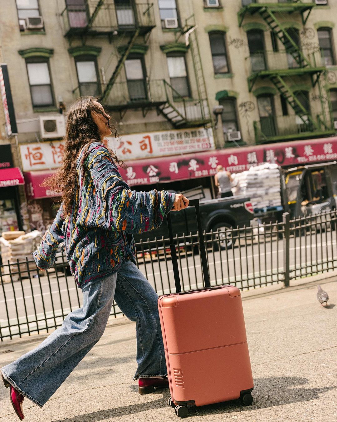 Adult woman with suitcase walks down urban street with apartment buildings and Chinese restaurant in background on sunny day.
