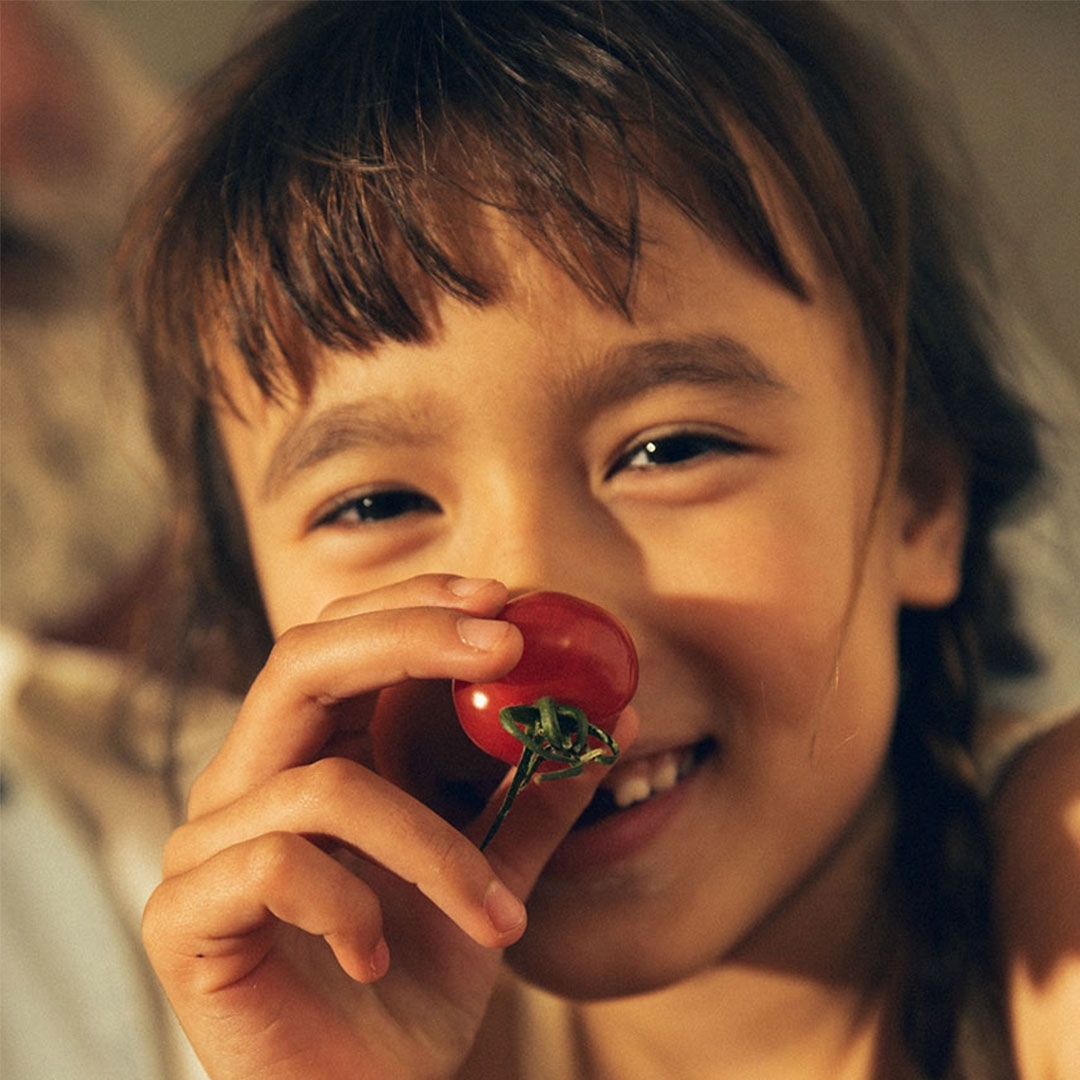 Smiling child holds red tomato in a close-up studio shot with soft, diffused lighting, showcasing natural beauty and fresh produce.
