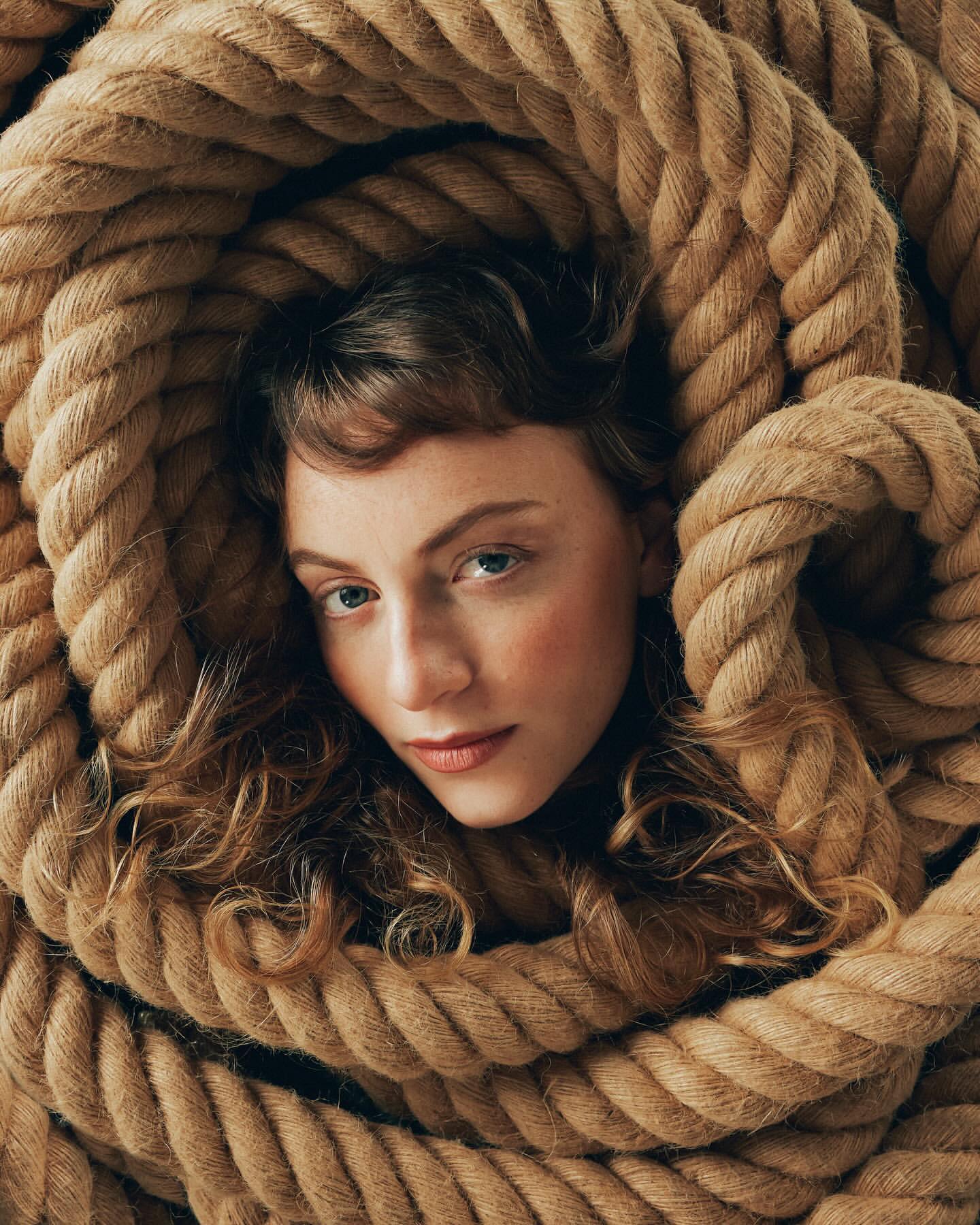 Eye-level portrait of an adult woman with curly hair, soft makeup, and neutral expression, framed by thick coils of tan rope.